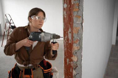 Kendine güvenen kadın güvenlik gözlükleri ve alet kemeri takan tamirci ev tadilatı sırasında eski ahşap kapıyı deliyor. Tamir, yeniden inşa, ev geliştirme kavramı.