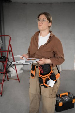Pano ve alet kemeri olan tamirci kadın yenileme odasında kırmızı merdivene ve alet çantasına karşı. Ev onarımı, organizasyon ve gelişimi somutlaştırır.