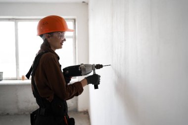 İnşaat ve tadilat sırasında duvarda matkapla delik açan gözlük takan genç bir kadının yan görüntüsü. Boşluğu kopyala