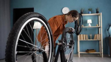 Gözlüklü genç kadın bisiklet operasyonunu kontrol ediyor. Dönen tekerlek ve elleri odada pedal çeviriyor. Boşluğu kopyala