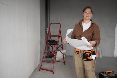 Tamirci kadın, inşaat sahasında ya da tadilat sırasında elinde kağıt planlar tutarken kemer takıyordu. DIY onarım, planlama ve inşa süreci kavramı. Boşluğu kopyala