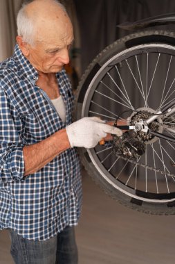 Evde ingiliz anahtarıyla bisiklet tekerleğini tamir eden yaşlı bir adamın portresi. DIY döngü onarımı, kapalı mekanik, ulaşım bakımı ve olgun yaşam tarzı aktivitesi kavramı. Boşluğu kopyala