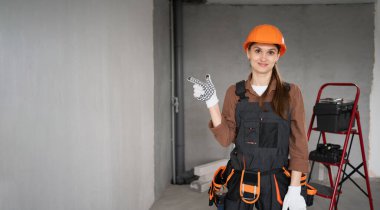 Kadın müteahhit portresi sabit şapka takıyor ve işçi tulumları fotokopi alanını işaret ediyor. İnşaat alanında parmak hareketiyle tamirci. Reklam, inşaat ve alışveriş konsepti. afiş, boşluğu kopyala