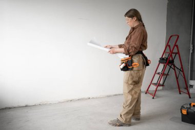Tamirci kadın, inşaat sahasında ya da tadilat sırasında elinde kağıt planlar tutarken kemer takıyordu. DIY onarım, planlama ve inşa süreci kavramı. Boşluğu kopyala