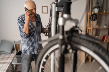 Emekli adam evde bisiklet tamir ediyor, tamirciye danışmak için telefon kullanıyor. Ulaşım onarımı kavramı, mekanik servis. Boşluğu kopyala