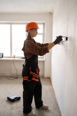 Kadın mühendis ya da mimar koruyucu bir miğferin duvar ölçüsünü ölçer. Evdeki bir inşaatçı beton bir duvarı kontrol ediyor. Onarım ve inşaat konsepti.