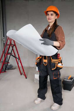 Tamirci kadın, tadilattan önce çıplak beton duvarlarla dolu odada elinde planlar tutuyor. İnşaat öncesi planlama, ev onarımı, iç mimari tasarım, DIY projesi.