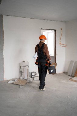 Alet kemerindeki kadın inşaatçı, bina yenilenmeden önce elinde planlar ve alet çantası tutuyordu. Arka plan