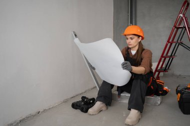 Genç bayan tamirci, işe başlamadan önce yenileme planı üzerinde çalışıyor, tasarım detaylarına odaklanıyor, ev geliştirme projesine hazırlanıyor..