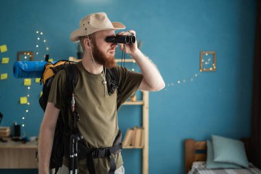 ... şapkalı, sırt çantalı bir erkek gezgin. Evde durup dürbünle bakıyor. Dağ yürüyüşü macerasına hazırlanıyor.