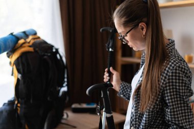 Kadın yürüyüşçü, dağ yürüyüşü için sırt çantası hazırlıyor. Elinde de evde kuzey yürüyüş direkleri var. Boşluğu kopyala