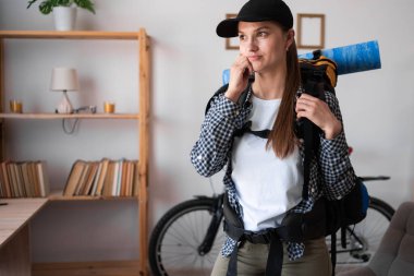 Düşünceli bir kadın sırt çantasıyla yürüyüşe çıkıyor. Evin oturma odasında dikilirken başka tarafa bakıyor. Boşluğu kopyala