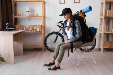 Sırt çantasıyla gezmeye hazır bir bayan yürüyüşçü oturma odasında oturmuş akıllı telefon kullanarak seyahat bilgilerini okuyor. Boşluğu kopyala