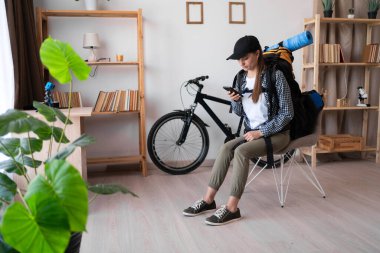 Sırt çantalı bayan gezgin oturma odasında cep telefonu kullanıyor, akıllı telefon uygulamasında sohbet ediyor, geziye çıkıyor. Boşluğu kopyala