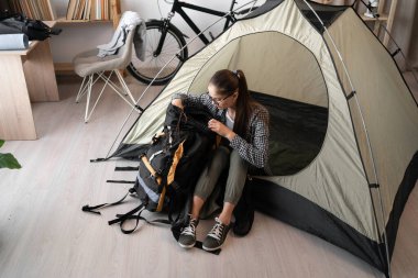 Gözlüklü ve günlük kıyafetleriyle sırt çantasıyla çadırda oturan sırt çantalı bir kadın eve dönüş yolculuğuna hazırlanıyor.