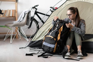 Gözlüklü ve günlük kıyafetleriyle sırt çantasıyla çadırda oturan sırt çantalı bir bayan geziye hazırlanıyor. Boşluğu kopyala