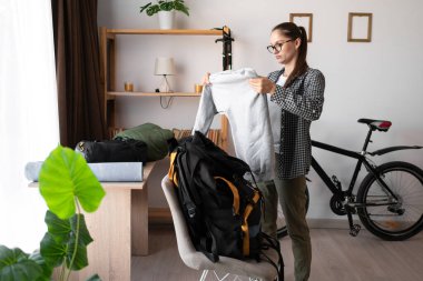 Tişörtlü, gözlüklü, sırt çantalı, teçhizatlı ve iç mekandaki gezisi için kıyafet taşıyan bir kadın. Seyahat, turizm ve yürüyüş konsepti