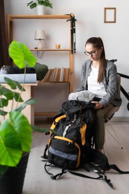 Tişörtlü, gözlüklü, sırt çantalı, teçhizatlı ve iç mekandaki gezisi için kıyafet taşıyan bir kadın. Seyahat, turizm ve yürüyüş konsepti