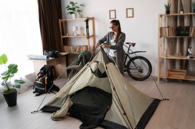 Evde dağ yürüyüşüne hazırlanan kadın yürüyüşçü, oturma odasında çadır kuran, yolculuktan önce kamp malzemelerini ve ekipmanlarını kontrol eden, açık hava macerası planlayan, yürüyüş hazırlığı kavramı.