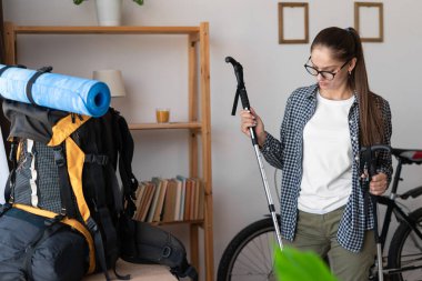 Kadın yürüyüşçü, dağ yürüyüşü için sırt çantası hazırlıyor. Elinde de evde kuzey yürüyüş direkleri var. Boşluğu kopyala