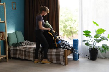 Woman packing Hiking backpack during preparing to trek in mountains. Camping equipment at home. Copy space