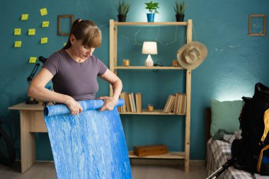 middle aged woman hiker rolling camping mat and packing hiking backpack at home, preparing for mountain journey, concept of tourism, outdoor travel, readiness and active lifestyle