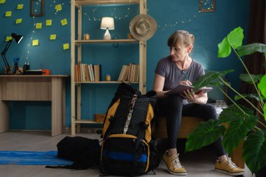 woman hiker sitting on floor writing checklist in notebook planning mountain trip at home, preparing equipment and organizing travel essentials, tourism and trekking concept