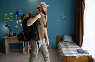 Sırt çantalı sakallı bir yürüyüşçünün portresi. Yürüyüş için hazırlanıyor. Evdeki bir odada duruyor.