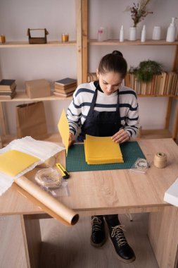 Önlüklü esmer esmer kadın elinde balmumu çarşaflarla atölyedeki masada oturan doğal balmumu çarşaflardan mum yapıyor. Boşluğu kopyala