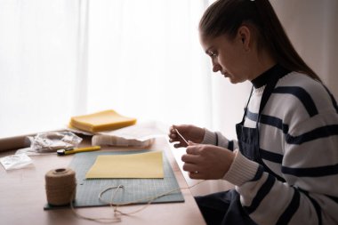 Önlüklü İspanyol esmer kadın balmumu çarşaflara ip koyup atölyedeki doğal balmumu çarşaflarından mum yapıyor. Uzayı kopyala Yan görünüm