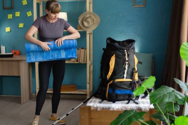 Evde büyük bir sırt çantası hazırlarken, bir kadın yolculuk sırasında uyumak için naylon paspas yuvarlıyor. Boşluğu kopyala