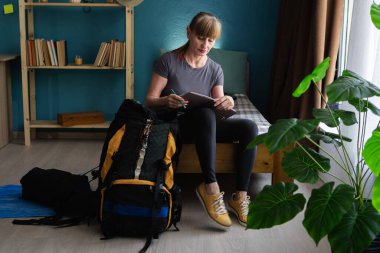 Üst düzey bir kadın bir kontrol listesi ile seyahat planlıyor sırt çantası için hazırlık yapıyor. Seyahat, planlama ve yürüyüş konsepti