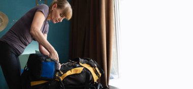 Evdeki yatakta sırt çantasına uyku tulumu ve farklı kamp malzemeleri koyan bir kadın. Uzayı kopyala Pankart