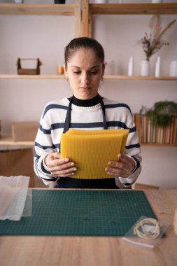 Önlüklü esmer kadın elinde balmumu çarşaflarla atölyedeki doğal balmumu çarşaflarından mum yapıyor. Boşluğu kopyala