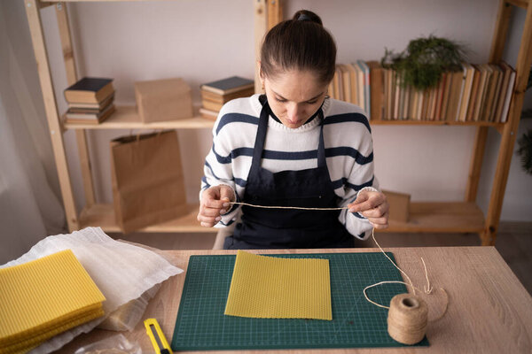 Hispanic brunette craftswoman in apron puts rope in beeswax sheets making candles from natural wax sheet in workshop. Copy space.