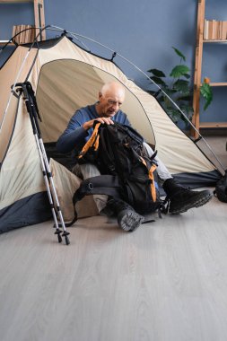 Yaşlı emekli adam içeride dağlara yürüyüş ve yürüyüş için hazırlanıyor, odada çadır kuruyor ve sırt çantalarını kontrol ediyor. Kıdemli gezgin evdeki kamp macerasına hazırlanıyor.