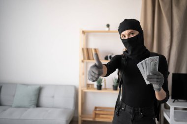 winner and lottery concept. female thief or robber in black balaclava robbing house showing class gesture and money bill into camera. close-up