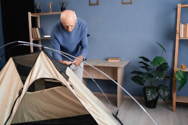 Kamp çadırı kuran bir emekli yürüyüş için hazırlanıyor ve ekipmanları evde kontrol ediyor. Boşluğu kopyala