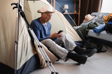 Emekli adam akıllı telefon kullanarak çadırda oturuyor evdeki dağ yürüyüşü ekipmanlarını kontrol etmeye hazırlanıyor. Boşluğu kopyala
