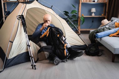 Yaşlı emekli adam içeride dağlara yürüyüş ve yürüyüş için hazırlanıyor, odada çadır kuruyor ve sırt çantalarını kontrol ediyor. Kıdemli gezgin evdeki kamp macerasına hazırlanıyor.
