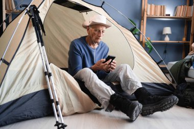 Emekli adam akıllı telefon kullanarak çadırda oturuyor evdeki dağ yürüyüşü ekipmanlarını kontrol etmeye hazırlanıyor. Boşluğu kopyala