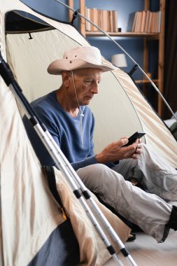 Emekli adam akıllı telefon kullanarak çadırda oturuyor evdeki dağ yürüyüşü ekipmanlarını kontrol etmeye hazırlanıyor. Boşluğu kopyala