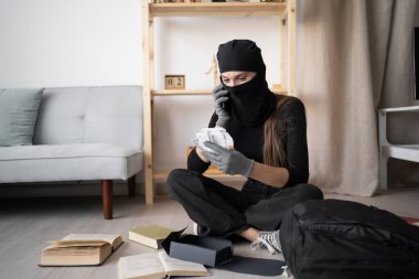 Siyah kar maskeli bir kadın hırsız ev telefonuyla suç ortaklarından para çalıyor ve yerde nakit parayla oturuyor. Boşluğu kopyala
