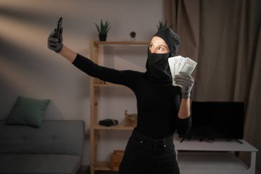 Hırsızlık ve soygun kavramı. Kadın hırsız ya da soyguncu siyah kar maskesi takıyor. Kameraya bakarak para çalarak selfie çekiyor.