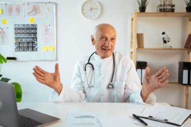 Steteskoplu olgun bir doktor dizüstü bilgisayarda çalışır ve kameraya bakar. Nazik bir gülümsemeyle el açar. Kıdemli terapist görüntülü arama yapıyor, pratisyen pratisyen pratisyen hekime danışmanlık yapıyor. Boşluğu kopyala