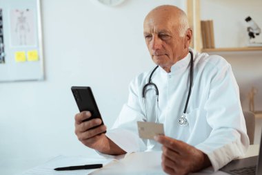 Yaşlı doktor akıllı telefon ve kredi kartını online ödeme için kullanıyor. Klinikte online bankacılık. Boşluğu kopyala