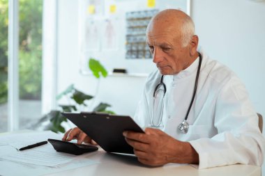 Elderly senior male Accounting doctor in white coat with stethoscope calculating bookkeeping finance and hospital budget in clinic interior, concept of healthcare and medicine