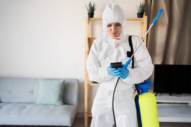 Haşere Kontrol Servisi 'nden kadın ilaçlayıcı sprey şişesiyle evde çalışıyor evde akıllı telefondan sohbet ediyor.