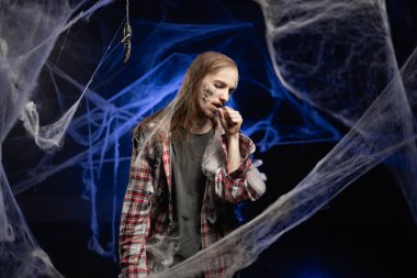 Creepy scary zombie man with ugly makeup cough having asthma attack, difficulties with breathe standing on spooky spider web background. Halloween concept. Horror film.