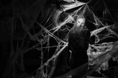 Creepy man with scary Halloween zombie makeup trapped spider webs, trying to scare. Dead guy in cob web on dark background. black and white photo. copy space